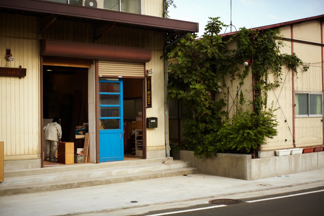The entrance to Akiyama Mokko furniture workshop in Yokohama, showing a cream and brown building with a blue door and ivy-covered walls.