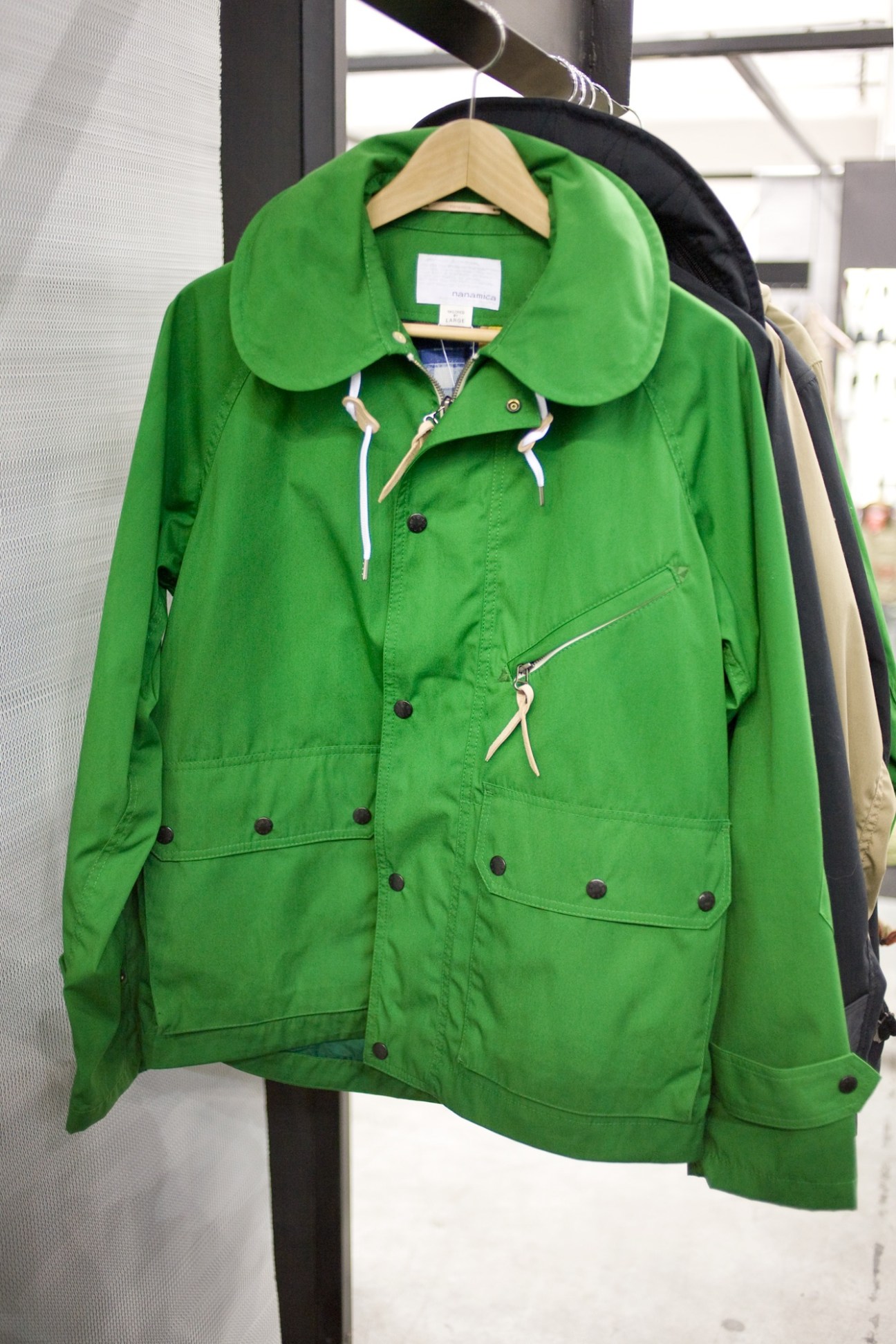 Bright green Nanamica jacket with oversized collar and white drawstring details on wooden hanger.