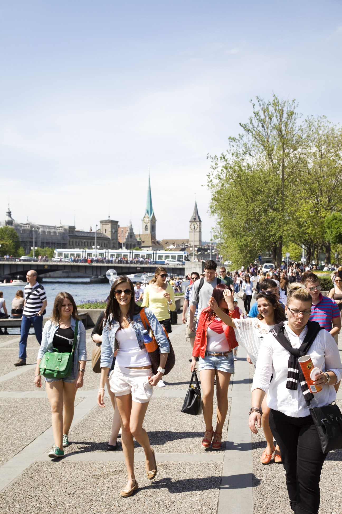 Crowds of people walking along a waterfront promenade in Zurich with churches and buildings visible across the water.