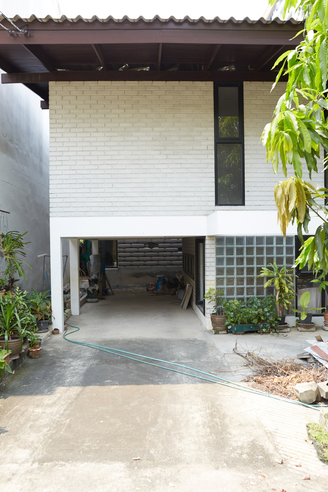 Two-story brick house renovation with white painted brick, dark metal roof overhang, and open ground-floor garage space surrounded by potted plants.