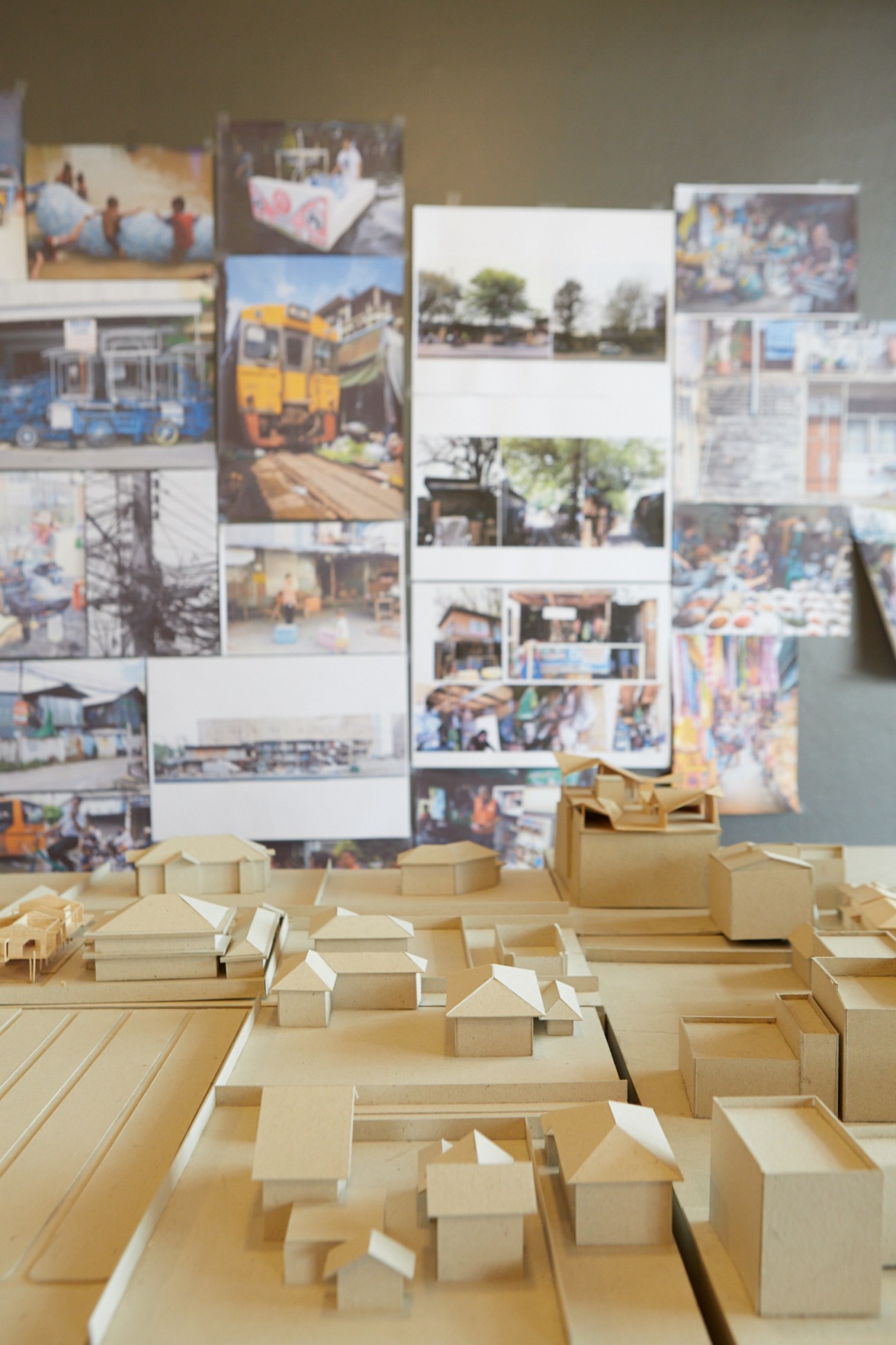 Wooden architectural scale models displayed on a table with reference photographs pinned to a wall behind them.