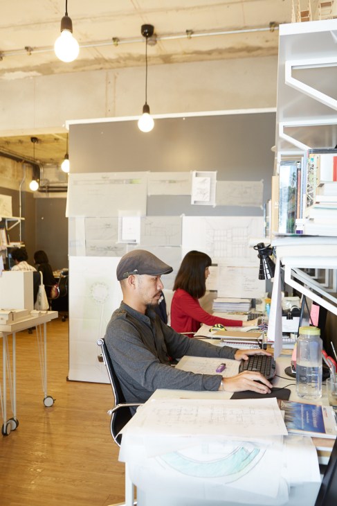 Two architects work at desks in a modern office with blueprint drawings and shelving visible.