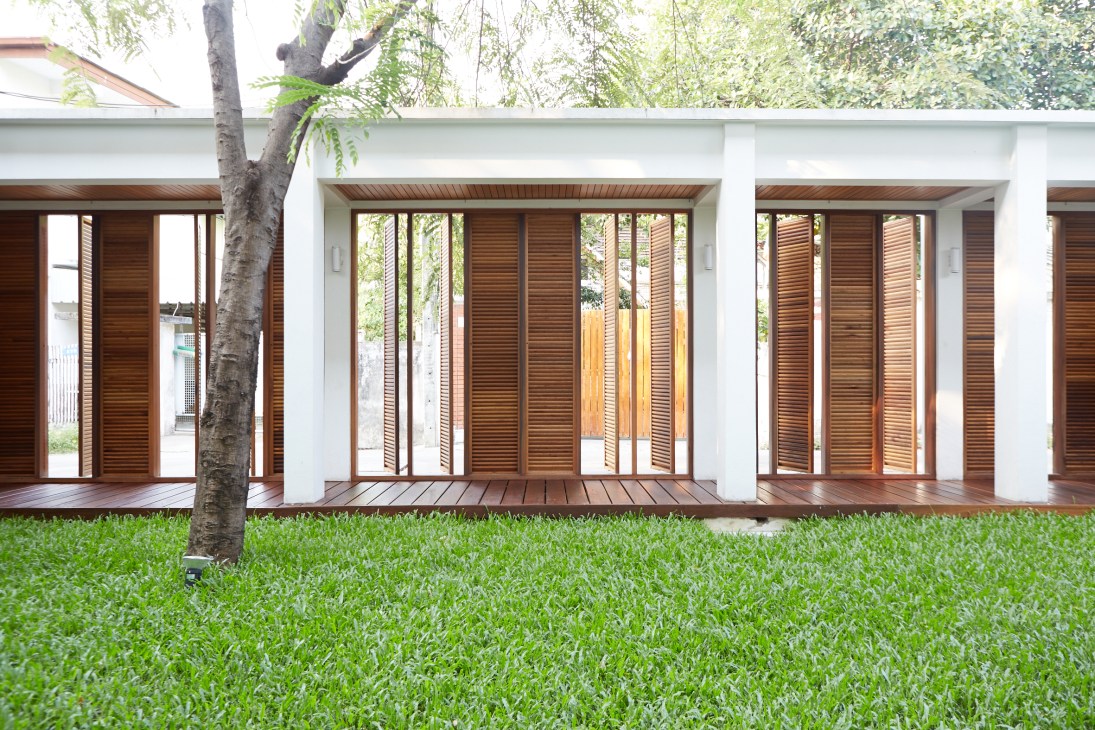Modern house with white walls and wooden louvered shutters opening onto a manicured lawn with a large tree.