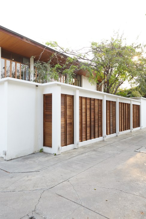 Modern white house facade with wooden shutters and upper-level windows, surrounded by green trees.