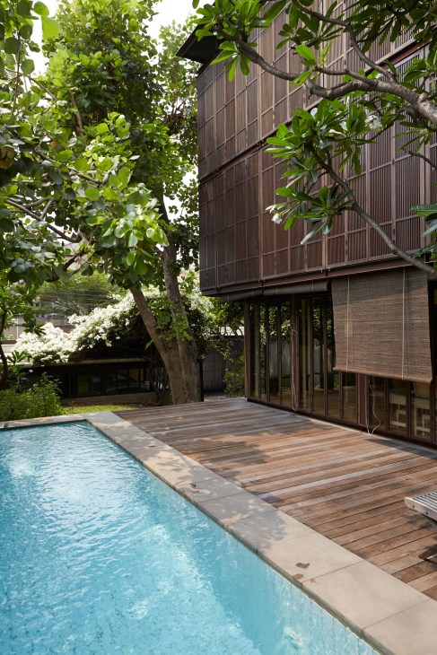 A modern residential building with metal screening elevated on concrete piles overlooks a swimming pool and wooden deck, surrounded by tropical vegetation.