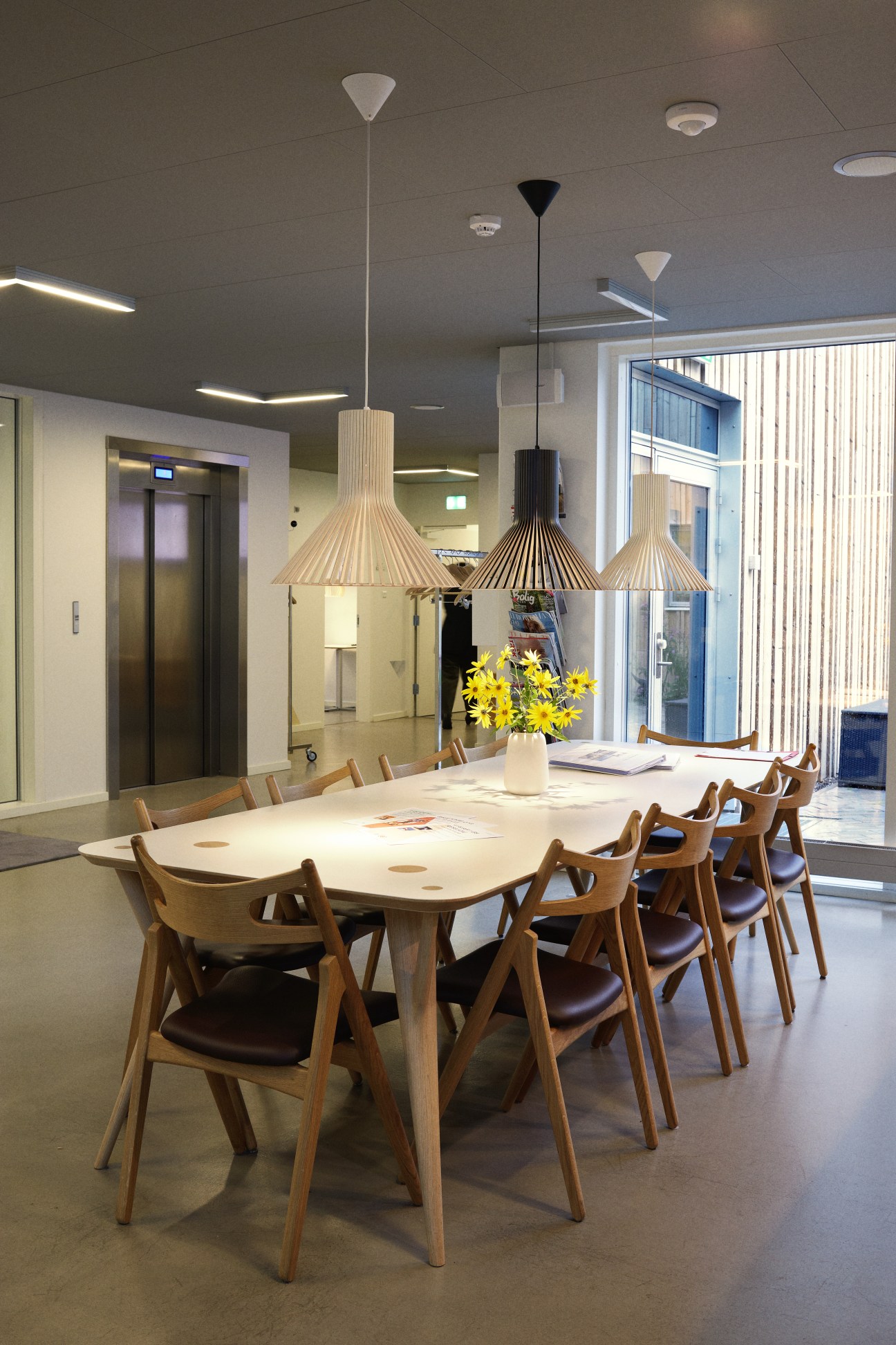 Modern café area with wooden chairs, pendant lights, and flowers on a long table.