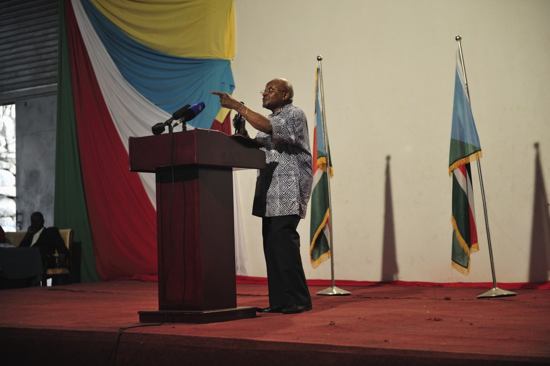 Archbishop Desmond Tutu speaking at a podium at the Nyakuron Cultural Centre in Juba, gesturing while delivering a speech, with flags in the background.