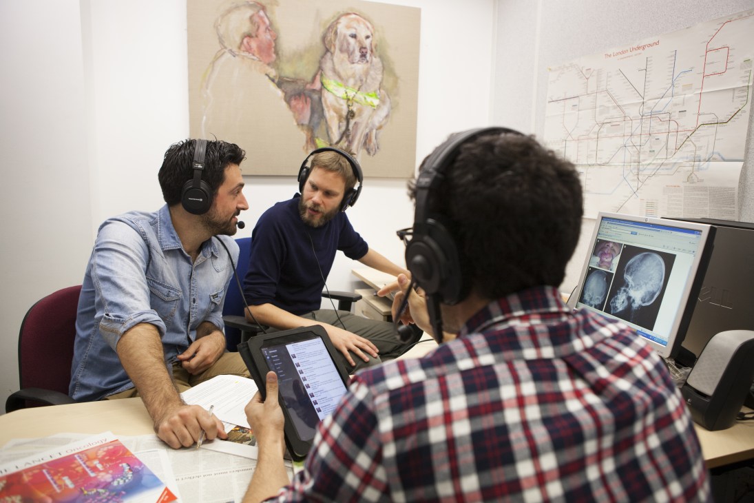 Three people wearing headphones record a podcast in an office with medical imaging visible on a monitor.