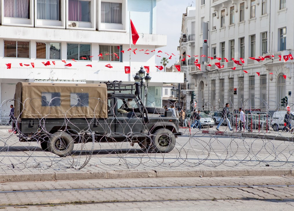Military truck surrounded by barbed wire on a Tunisian street with Tunisian flags flying above buildings.