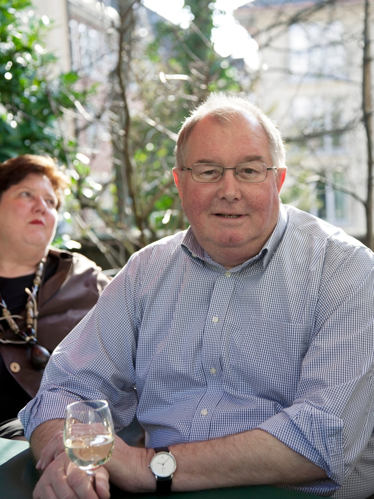 Thomas Weber sits outdoors with a glass of wine, wearing glasses and a blue checkered shirt.