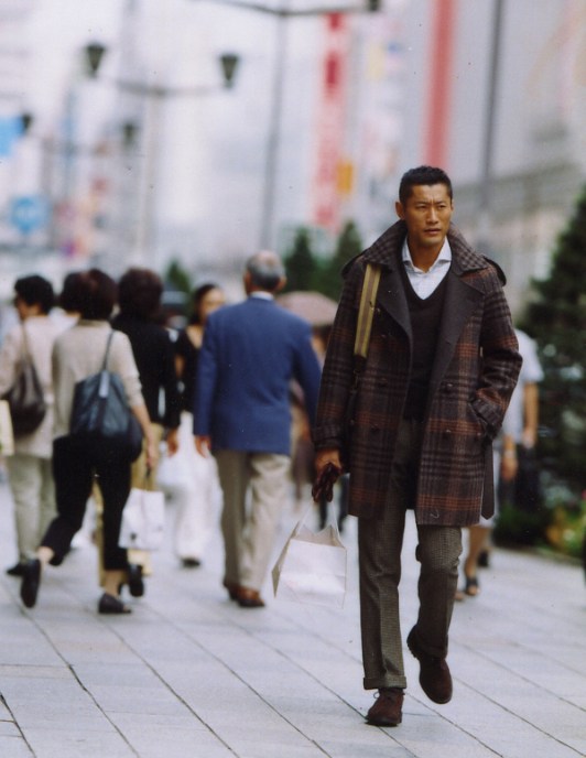 Man wearing plaid coat and tan scarf walking through Tokyo street among pedestrians.
