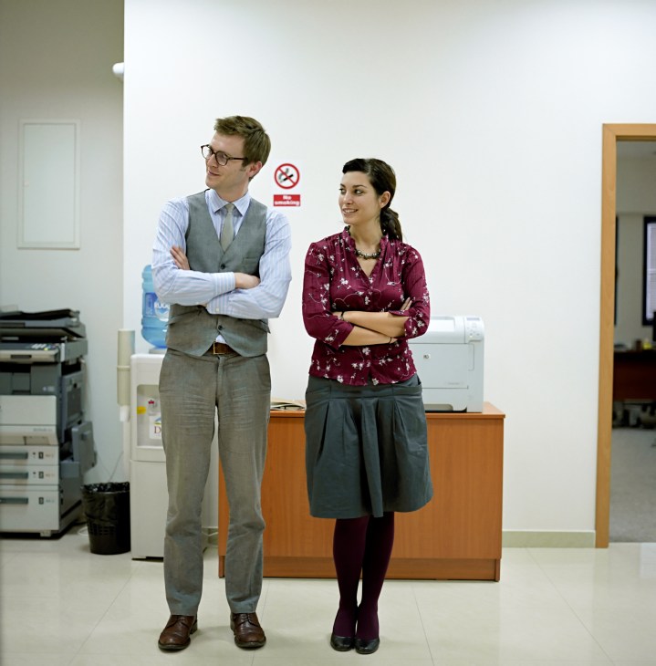 Two professionals stand in an office: a man in a grey vest and glasses, and a woman in a burgundy floral blouse.