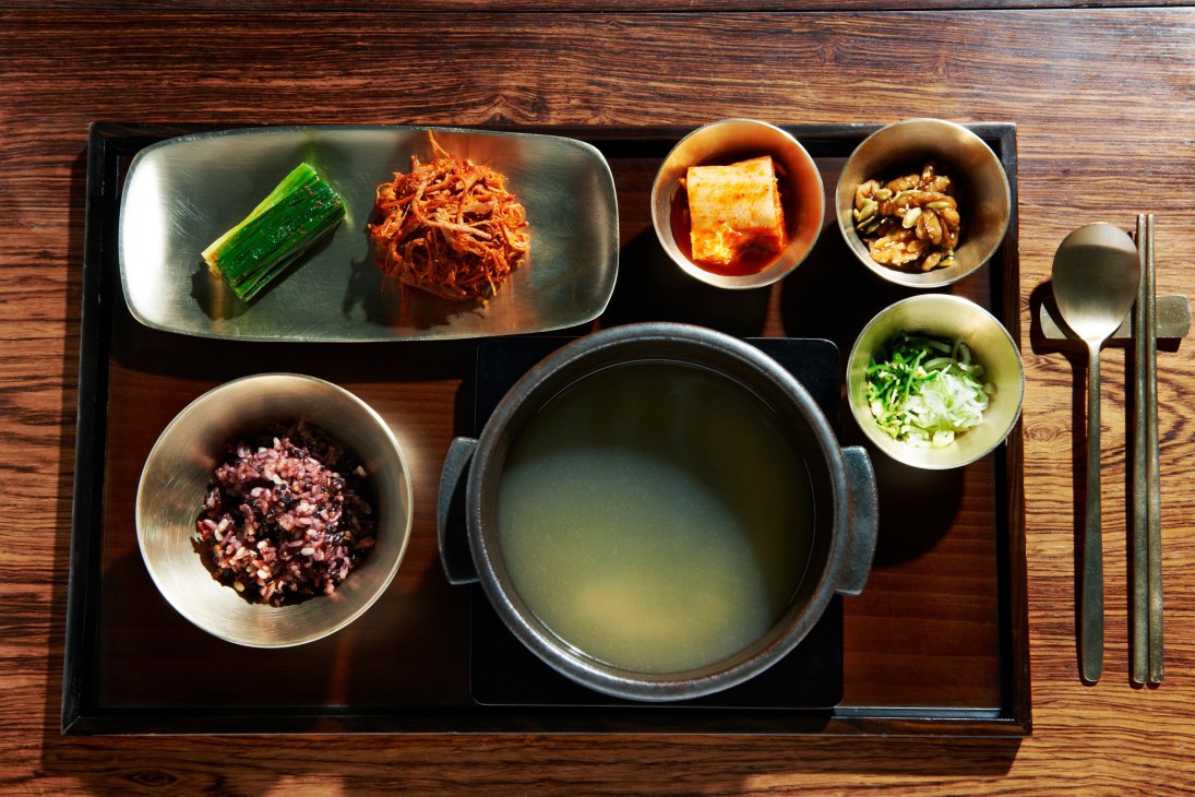 Korean fine dining meal at O-neul restaurant featuring a black tray with broth, vegetables, kimchi and banchan side dishes on wooden table.