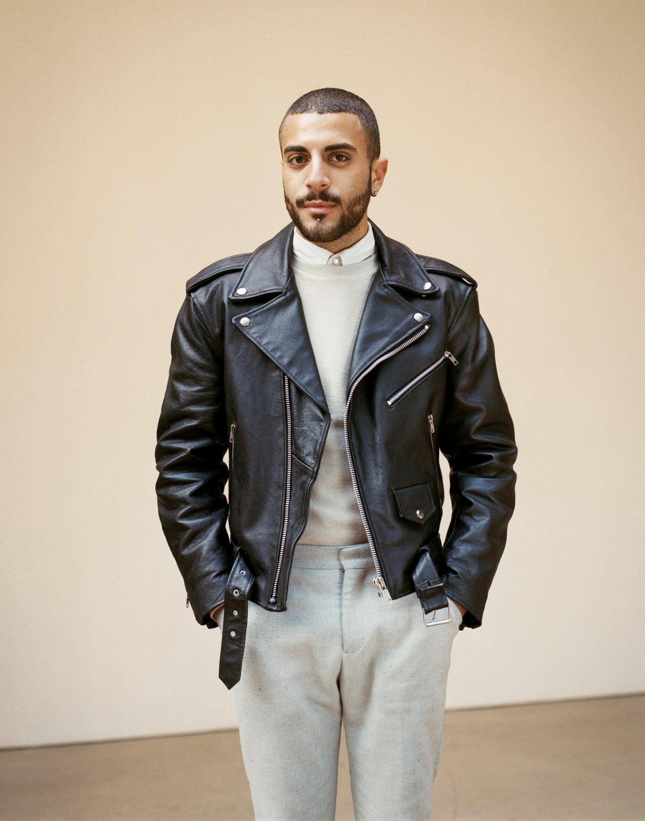 Man wearing black leather jacket and light-coloured trousers against a beige wall.