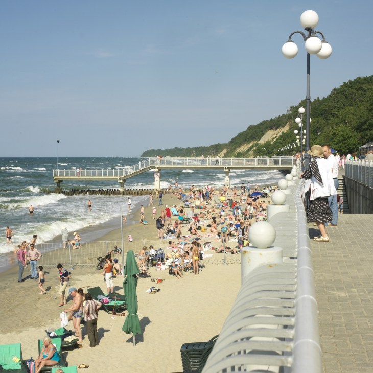 Crowded beach scene at Svetlogorsk with pier, lamp post, and forested cliffs in background.