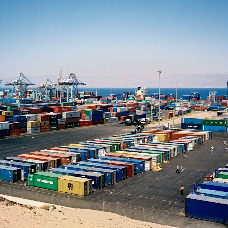 Shipping containers stacked at Aqaba port with cargo cranes and Red Sea in background.