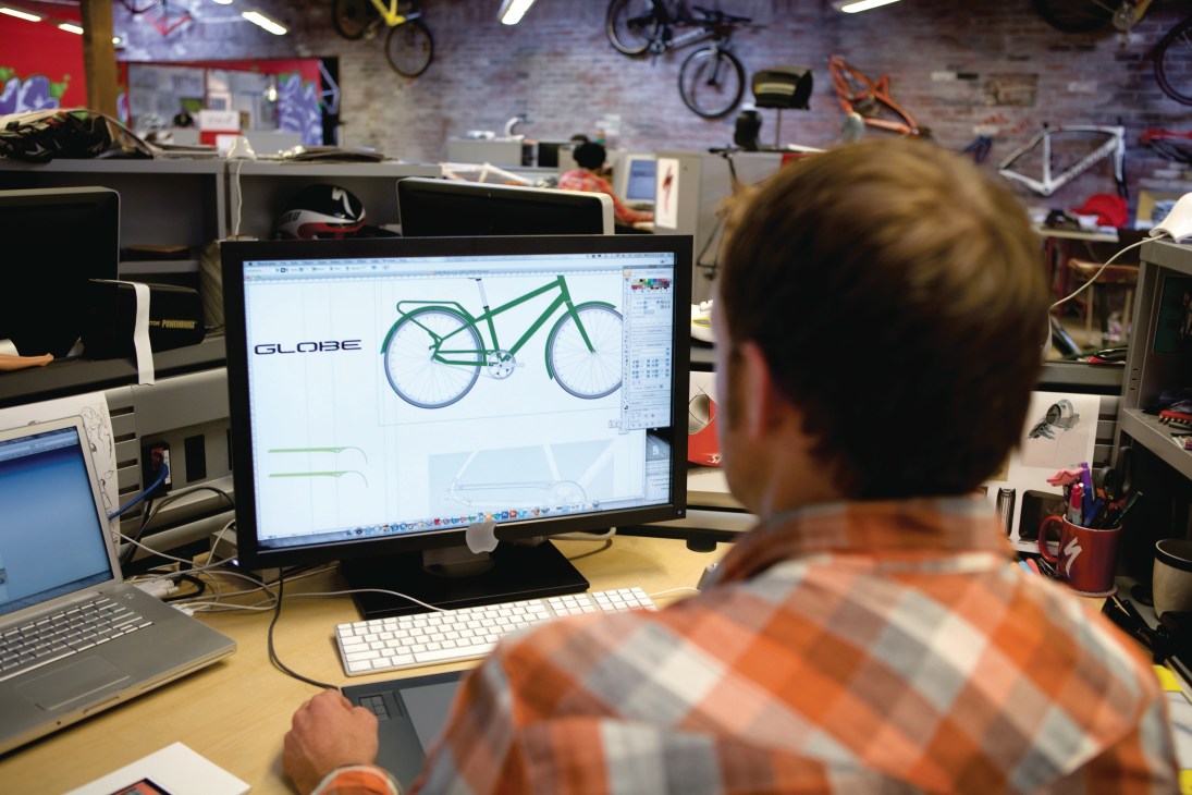 Globe bike design displayed on computer monitor in office workspace with bikes visible on wall.