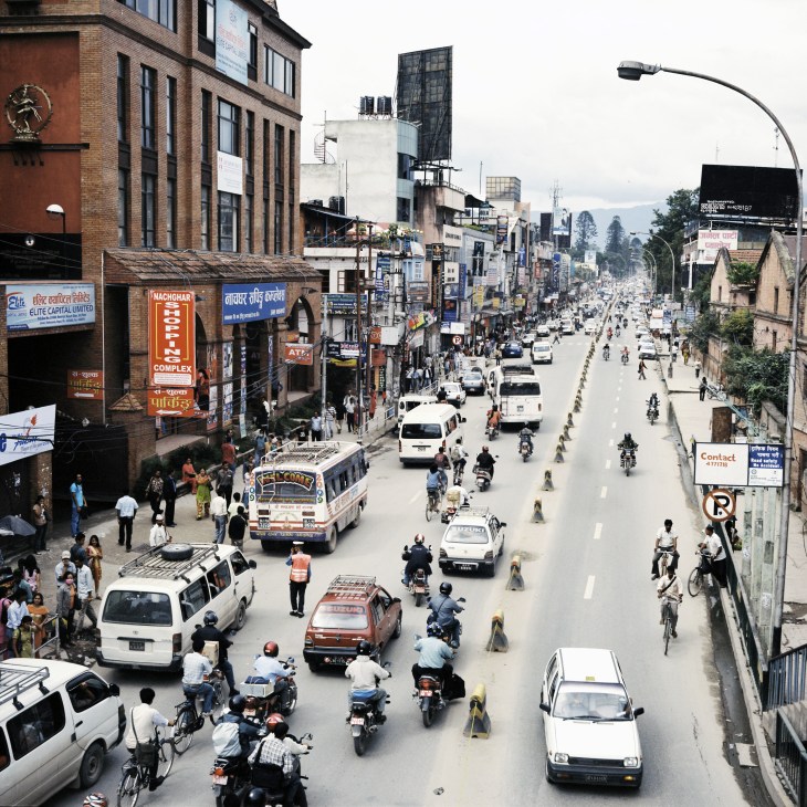 A busy street scene in downtown Kathmandu with vehicles, motorcycles, bicycles and pedestrians amid colorful shop signs in Nepali script.