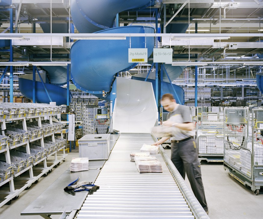 Worker stacking newspapers on a conveyor belt in an automated Swiss Post sorting facility with blue machinery overhead.