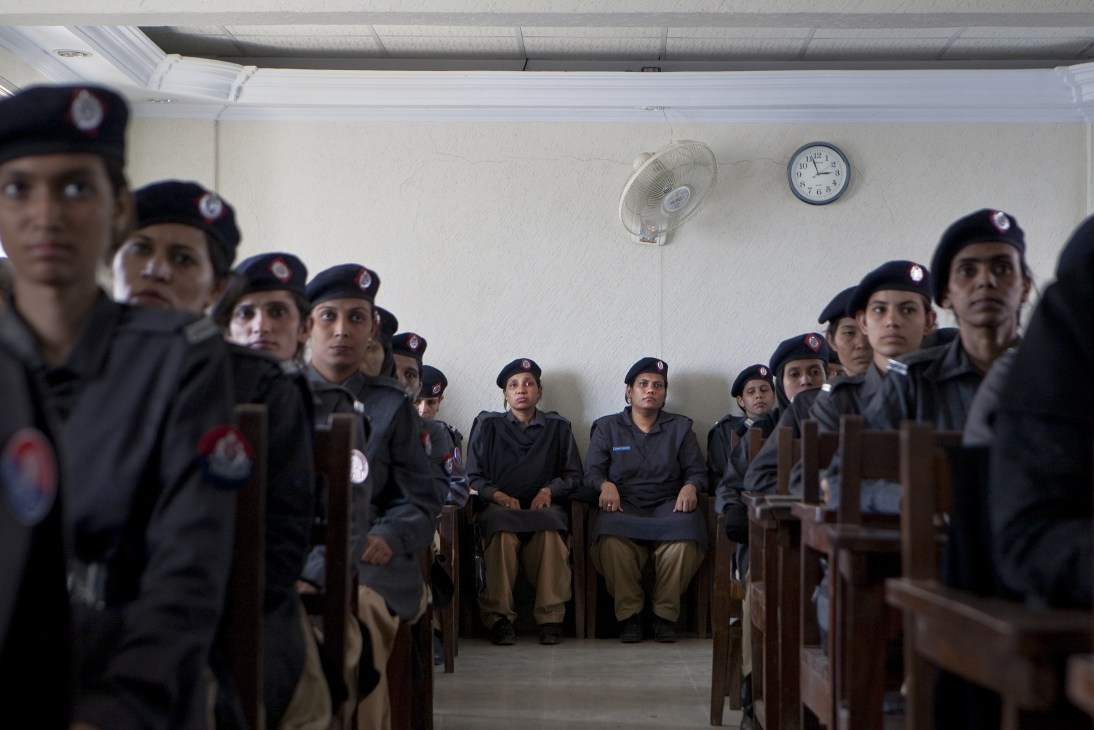 Female police trainees in dark uniforms and berets sit in a training room with a clock and fan on the wall.