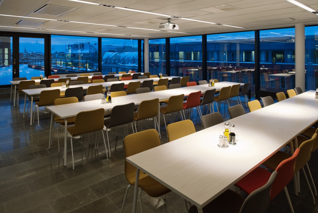 Modern restaurant and auditorium space with white tables, colorful chairs in gold, black and orange, and floor-to-ceiling windows overlooking Stockholm at dusk.