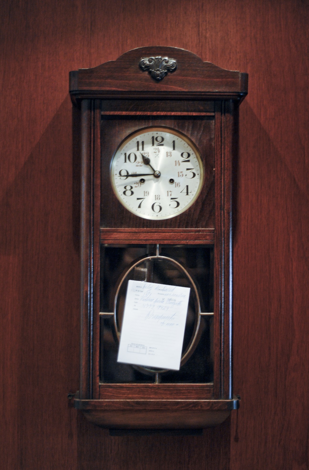 Wooden pendulum clock with white face ready for repair against a burgundy wall.