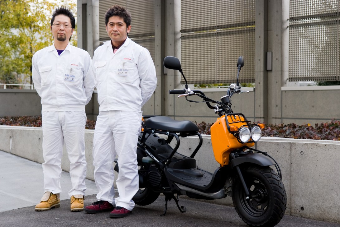 Two Honda engineers in white uniforms stand next to an orange and black Honda Zoomer scooter.