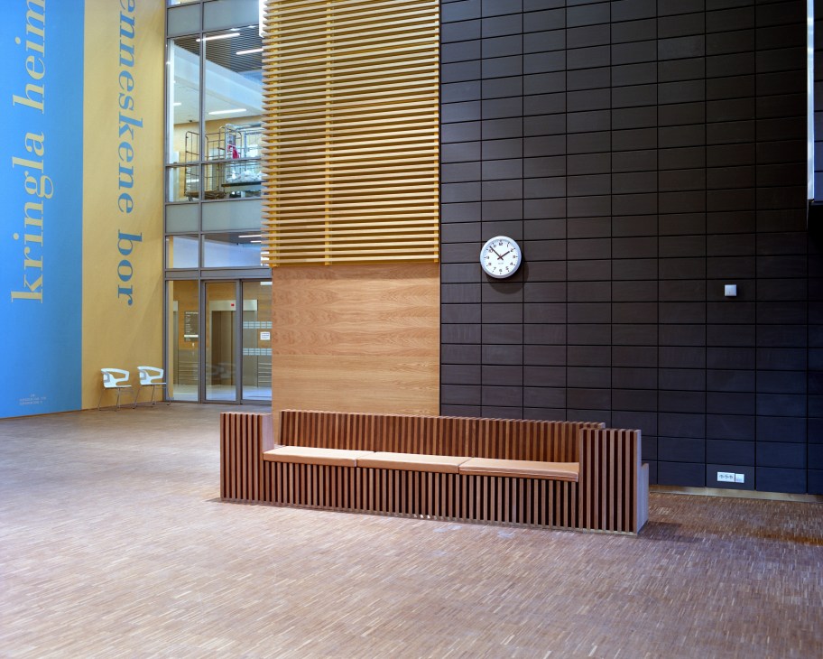 Hospital interior with wood-slat seating bench, blue signage, and clock on dark tile wall.