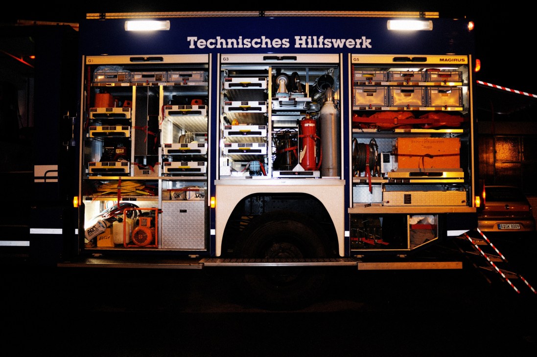 A THW rescue truck with open cargo doors displaying organized tools, machinery, fire extinguishers, and equipment in illuminated compartments.