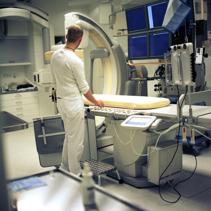 Medical technician operating an angiography apparatus for cardiac X-ray imaging in a hospital room.
