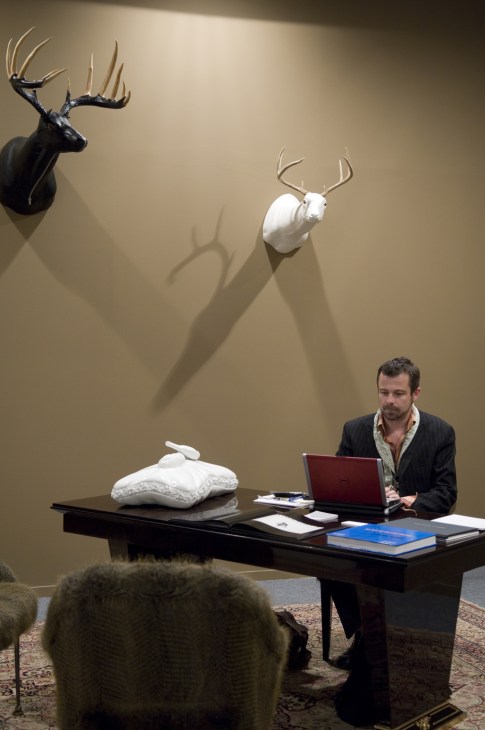 A man in a dark blazer works at a desk with a laptop, surrounded by taxidermied animal heads with antlers mounted on a beige wall above.