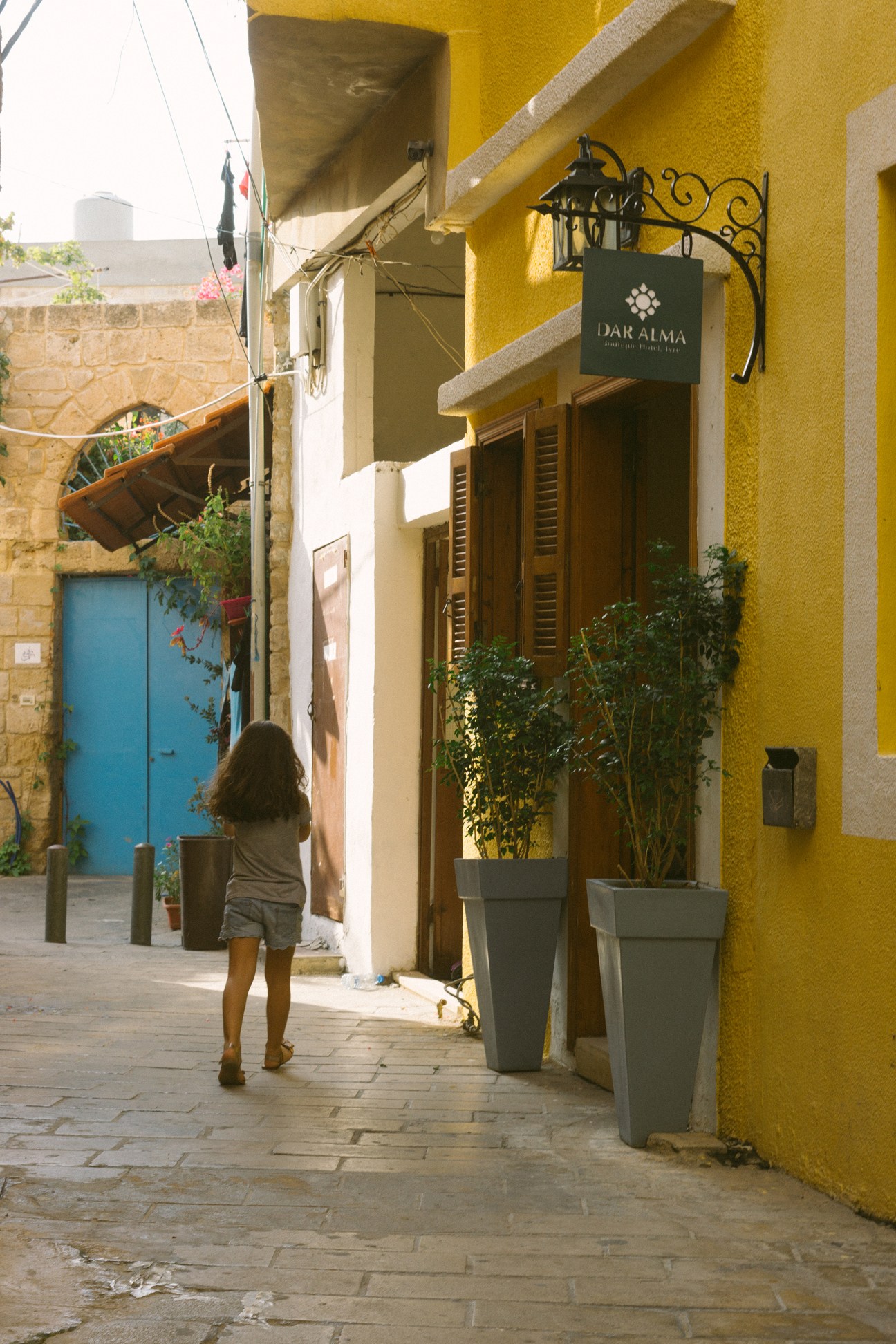 A child walks down a narrow cobblestone street past a bright yellow building with a green Dar Alma sign in Tyre's historic district.