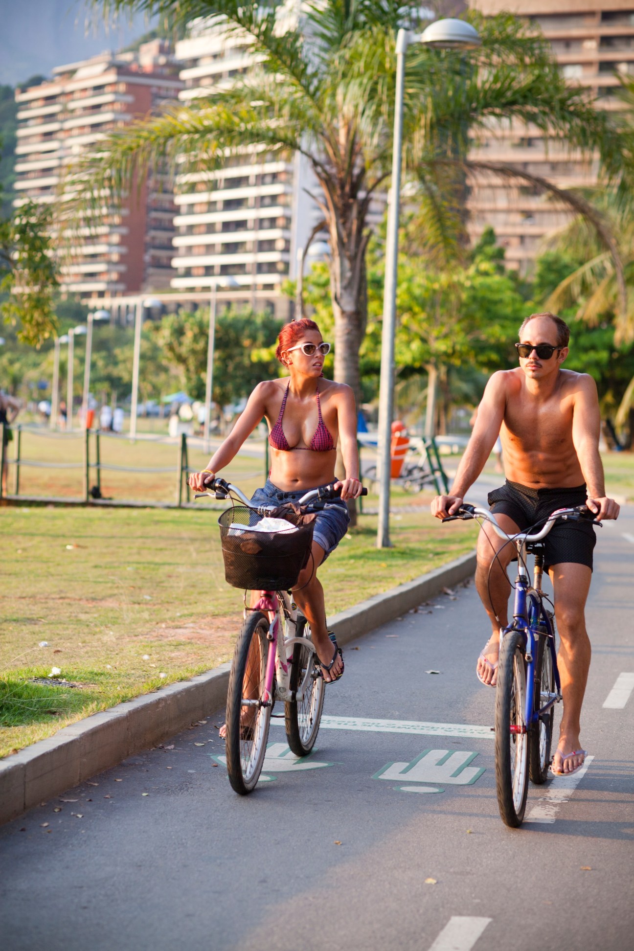 Two cyclists ride along a dedicated cycle lane with apartment buildings and palm trees in the background in Rio de Janeiro.
