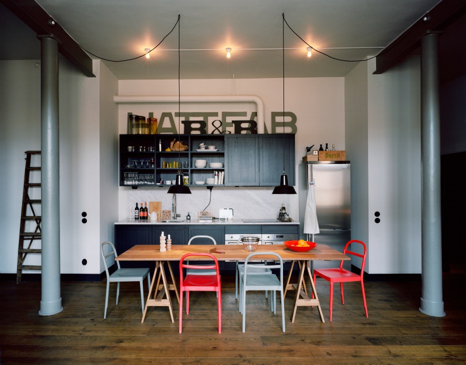 Modern kitchen and dining area with wooden table, colorful chairs, dark cabinetry, and pendant lights.