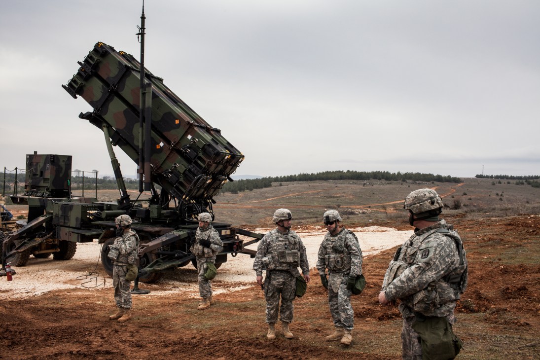 US military personnel stand near a truck-mounted PAC-2 Patriot air defense launcher angled skyward on scrubland.