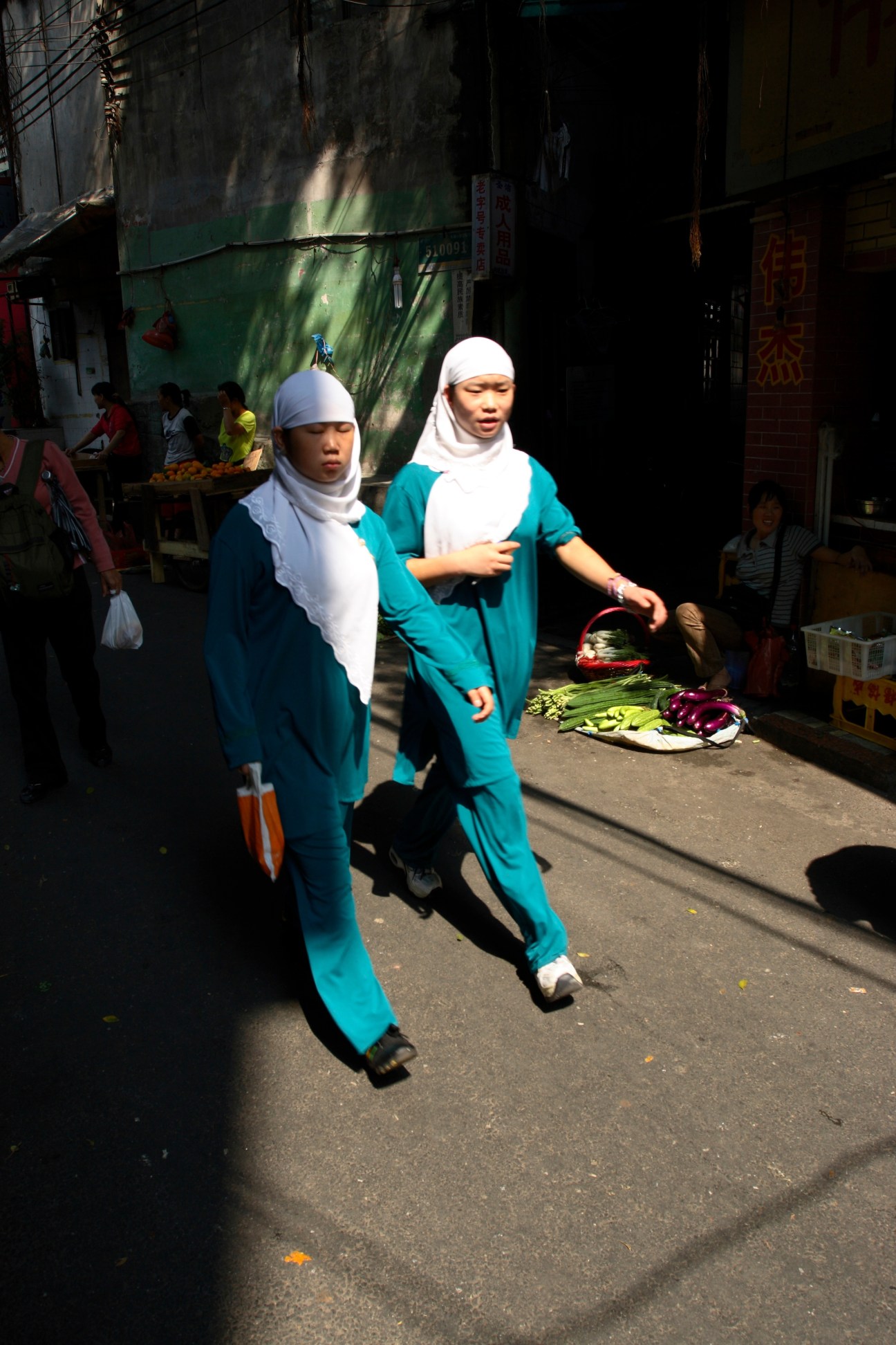 Two Muslim girls wearing headscarves and teal outfits walk through a Guangzhou street market.