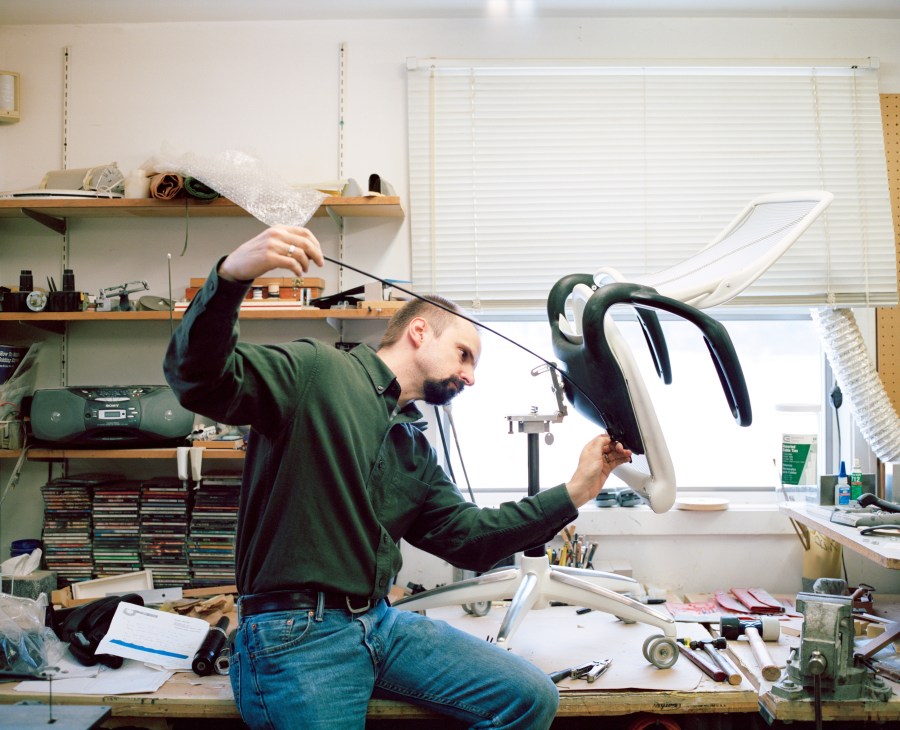 Jim Sorter examines a white chair prototype in a cluttered workshop filled with design tools and materials.