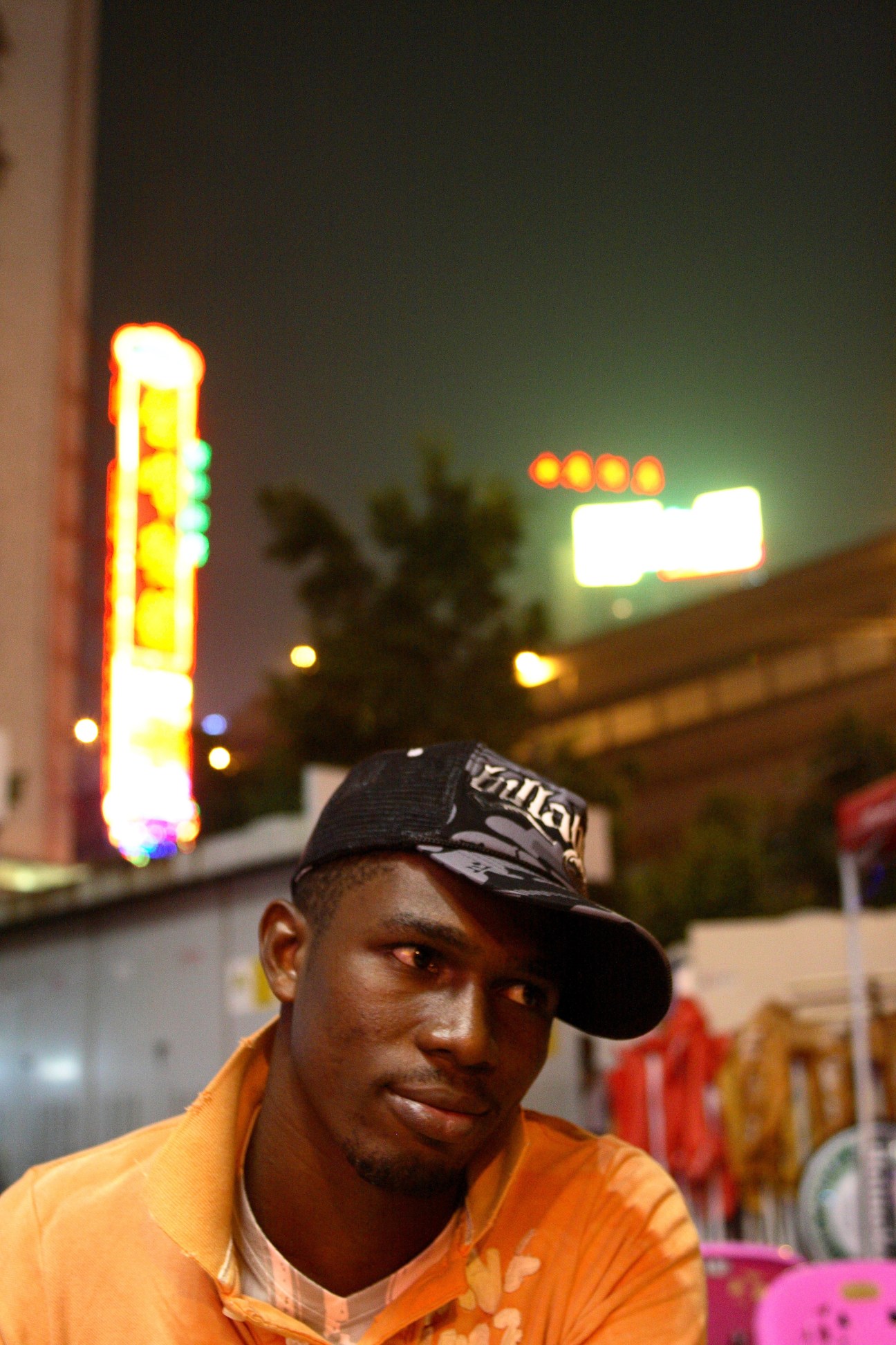 African man in orange shirt and black cap standing outside a bar at night with illuminated signs behind him.