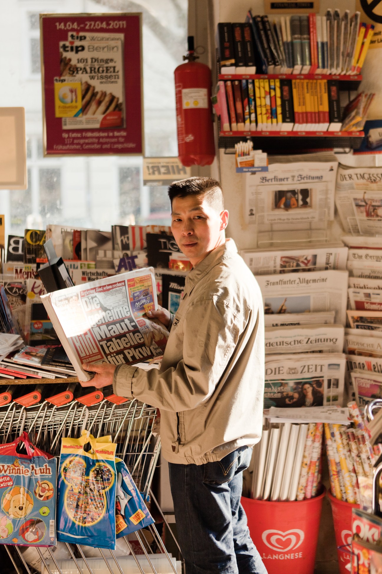 Van Dan Le holds a German newspaper while standing in his kiosk surrounded by publications and shelves of magazines.