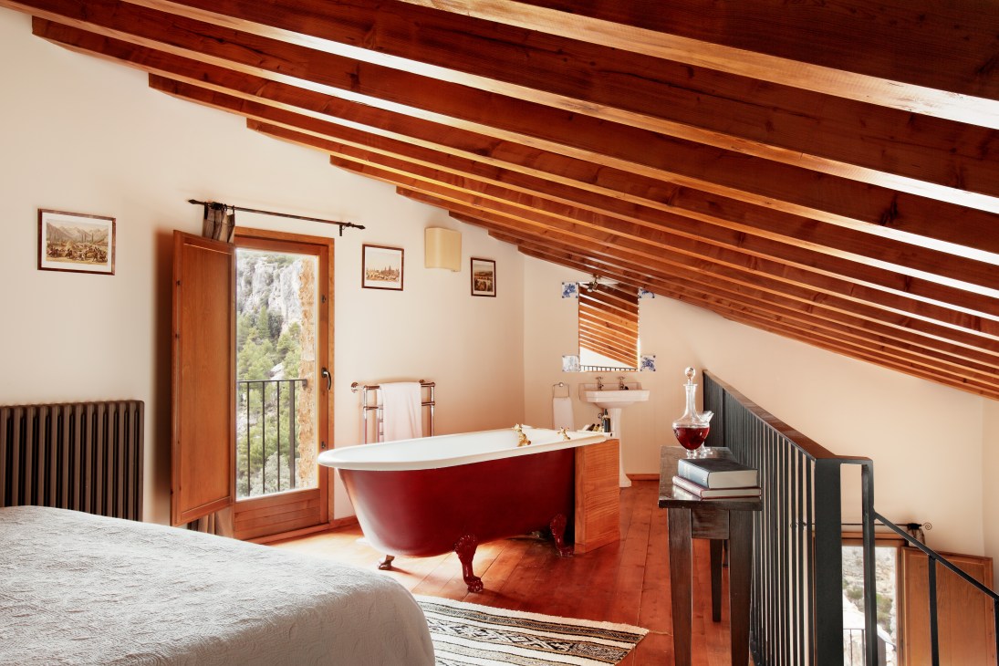 A rustic hotel bedroom with exposed wooden beams, a red freestanding bathtub, and views of countryside through French doors.