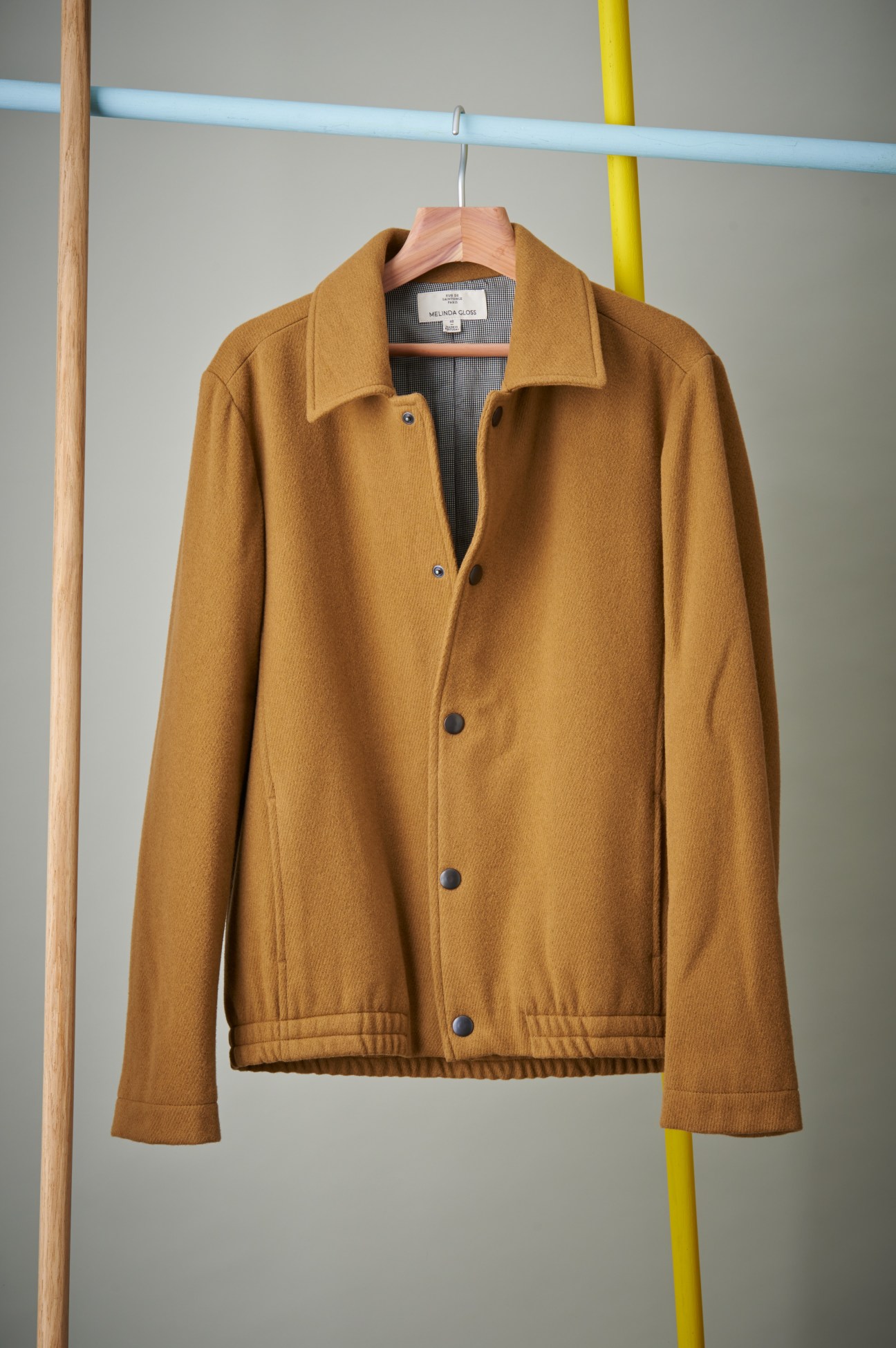 Camel-colored wool jacket by Melinda Gloss hanging on wooden hanger against gray wall with blue and yellow pipes.