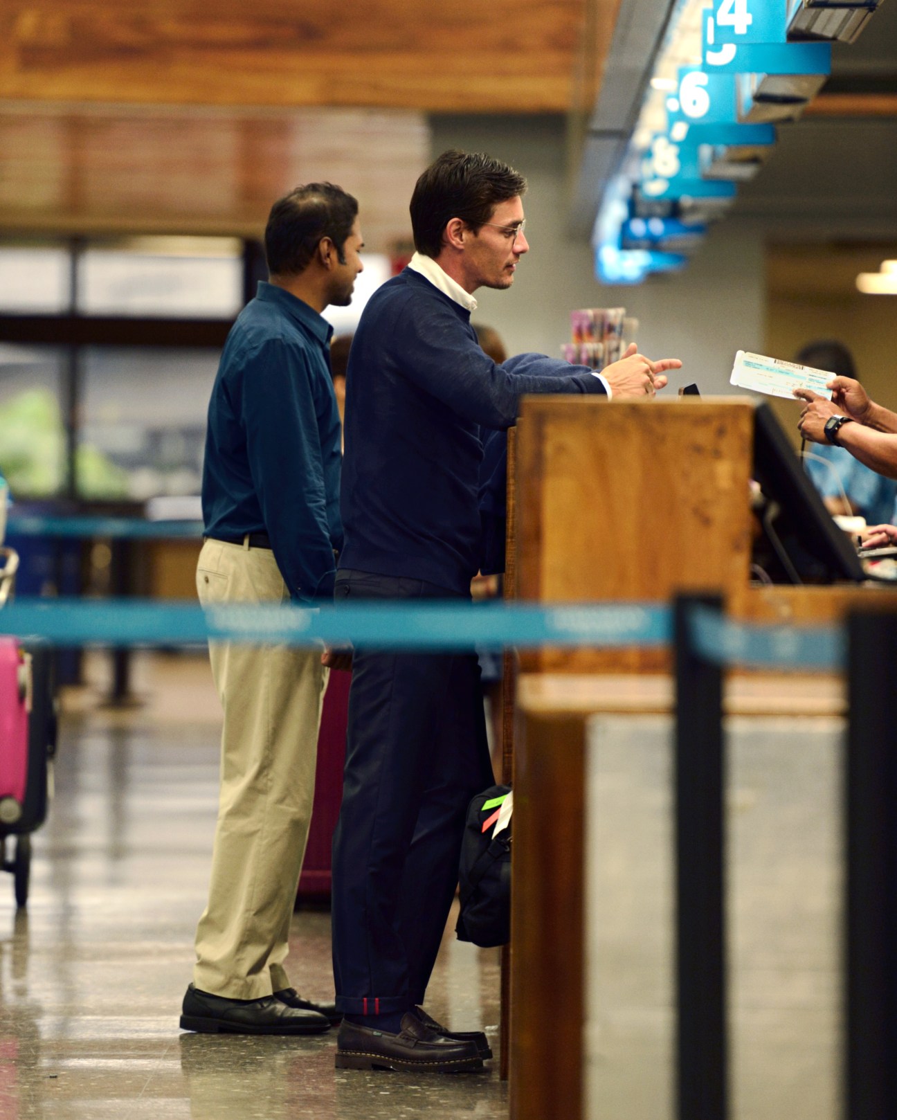Two men at an airport counter, one wearing a jumper and navy jacket with document case and bag.
