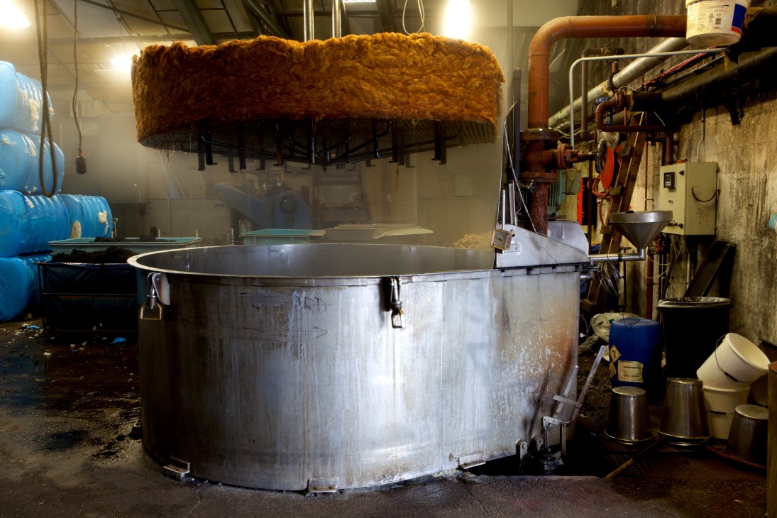 Large metal vat in a Harris Tweed mill with golden-brown dyed wool suspended above on an industrial wringing machine.