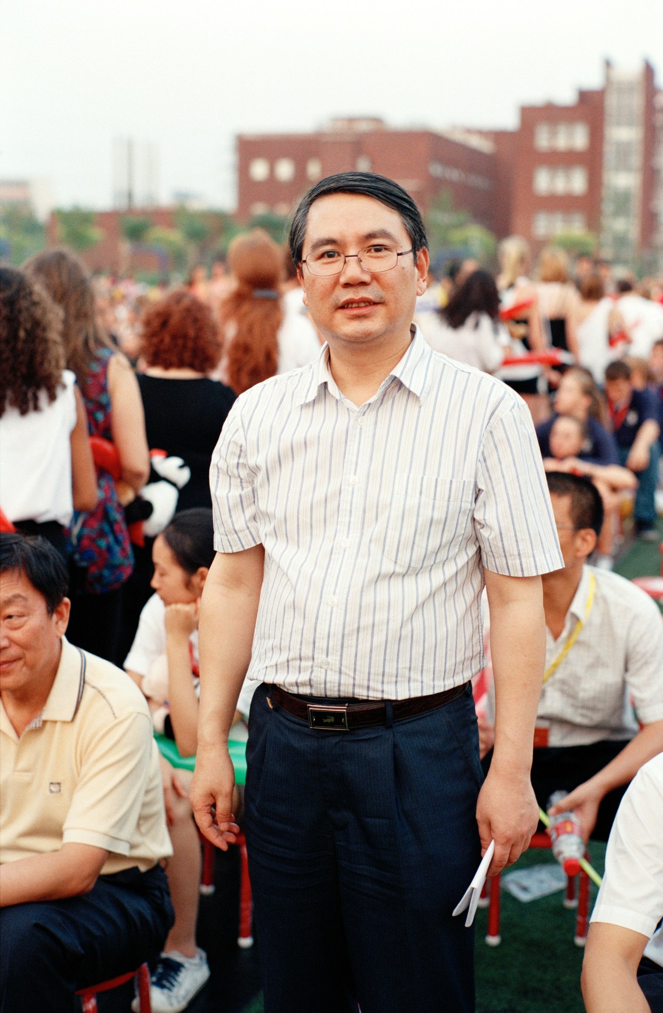 Wang Yongli, deputy director of Hanban, standing at the Chinese Bridge summer camp opening ceremony in Beijing with crowds and red brick buildings behind him.