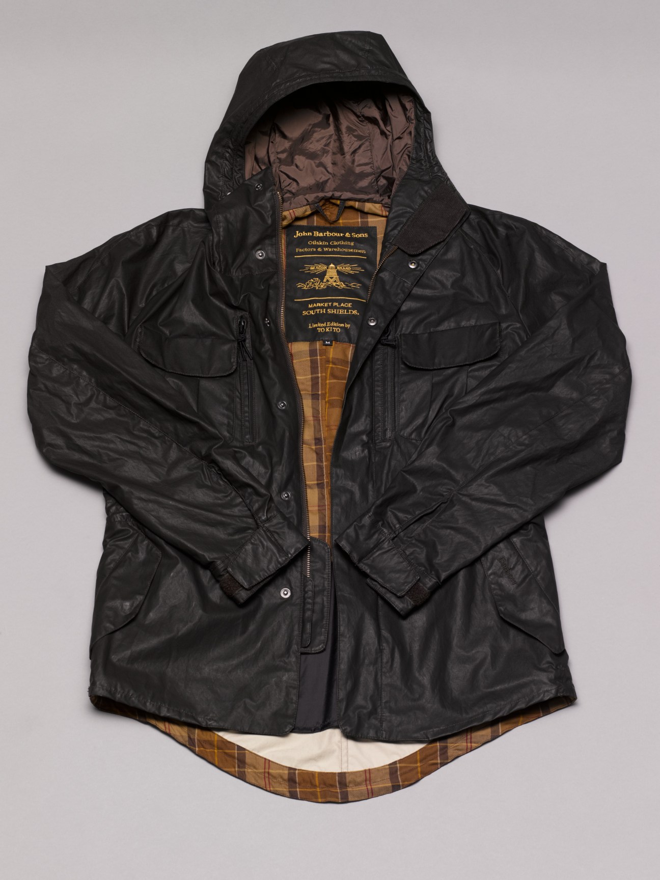Black wax Barbour jacket with gold tartan lining and hood displayed flat against white background.