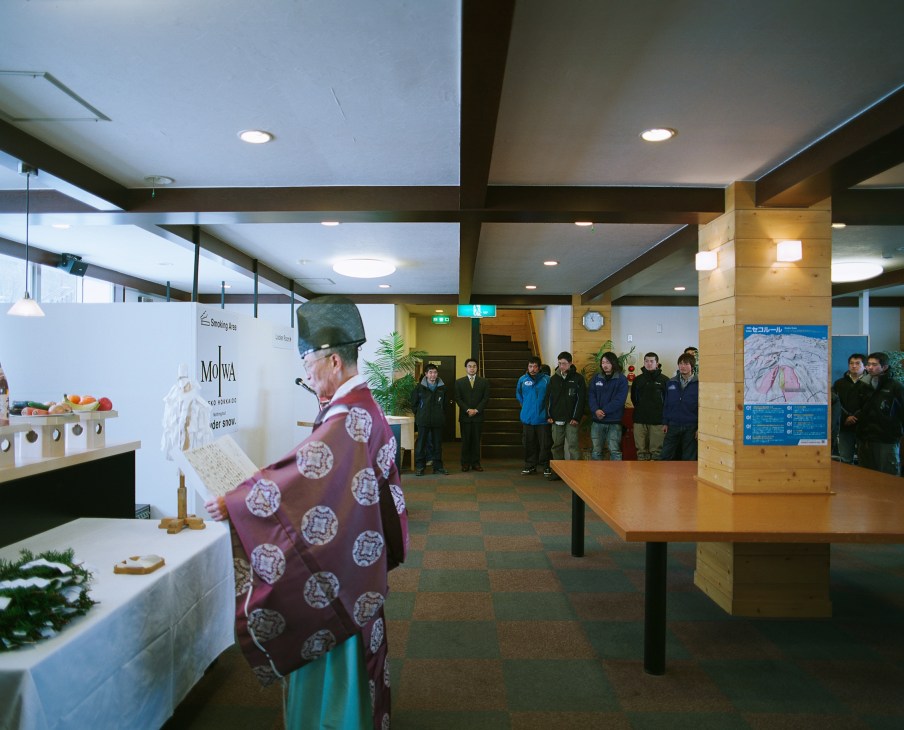 A Shinto priest in burgundy robes performs a blessing ceremony in a modern ski resort lobby with visitors gathered in the background.