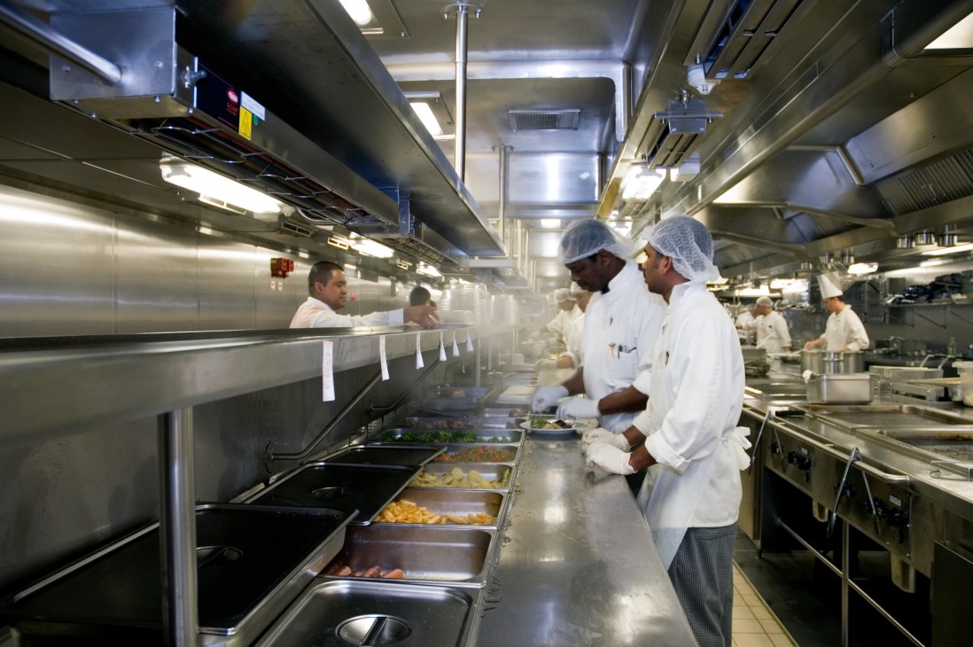 Chef and kitchen staff in white uniforms working in a professional ship galley with stainless steel equipment and food preparation stations.