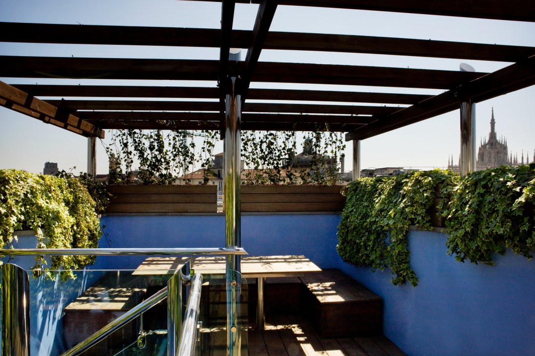 Teak observation deck with blue walls, climbing ivy, pergola and views of Milan cathedral and Alps.