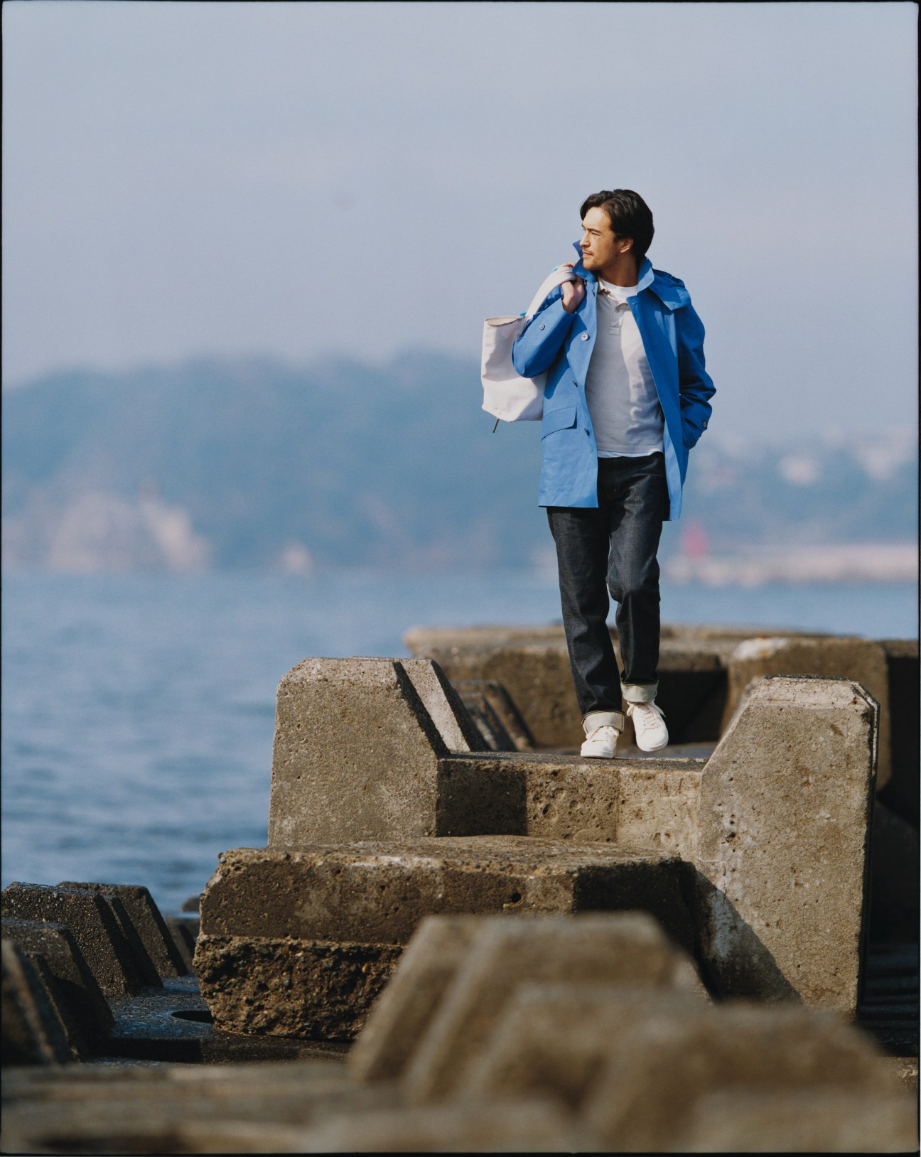 Male model wearing blue Gucci jacket and John Smedley polo shirt standing on concrete breakwater by the sea.