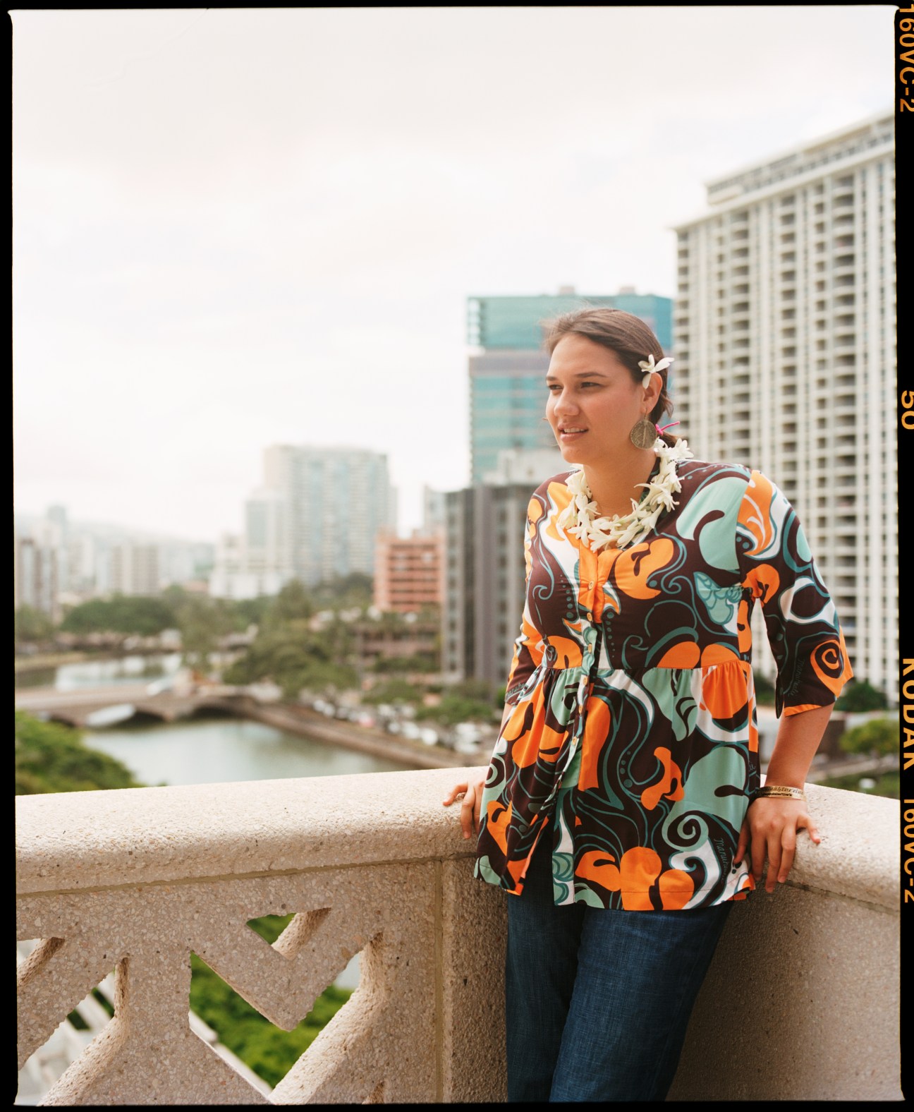Keli'i Wilson of Hawaiian Tourism stands on a balcony overlooking Waikiki, wearing a colorful patterned shirt and white flower in her hair.